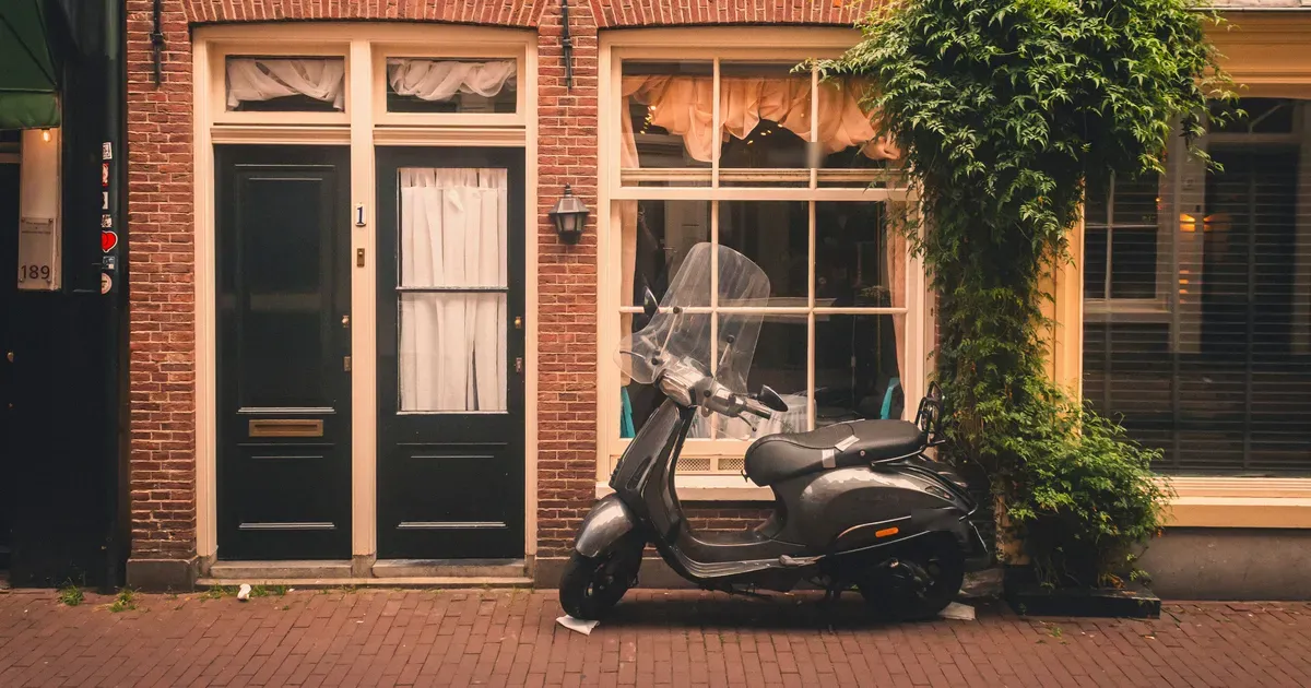 Grijze scooter geparkeerd voor een bakstenen pand met donkere deuren en een grote ruit